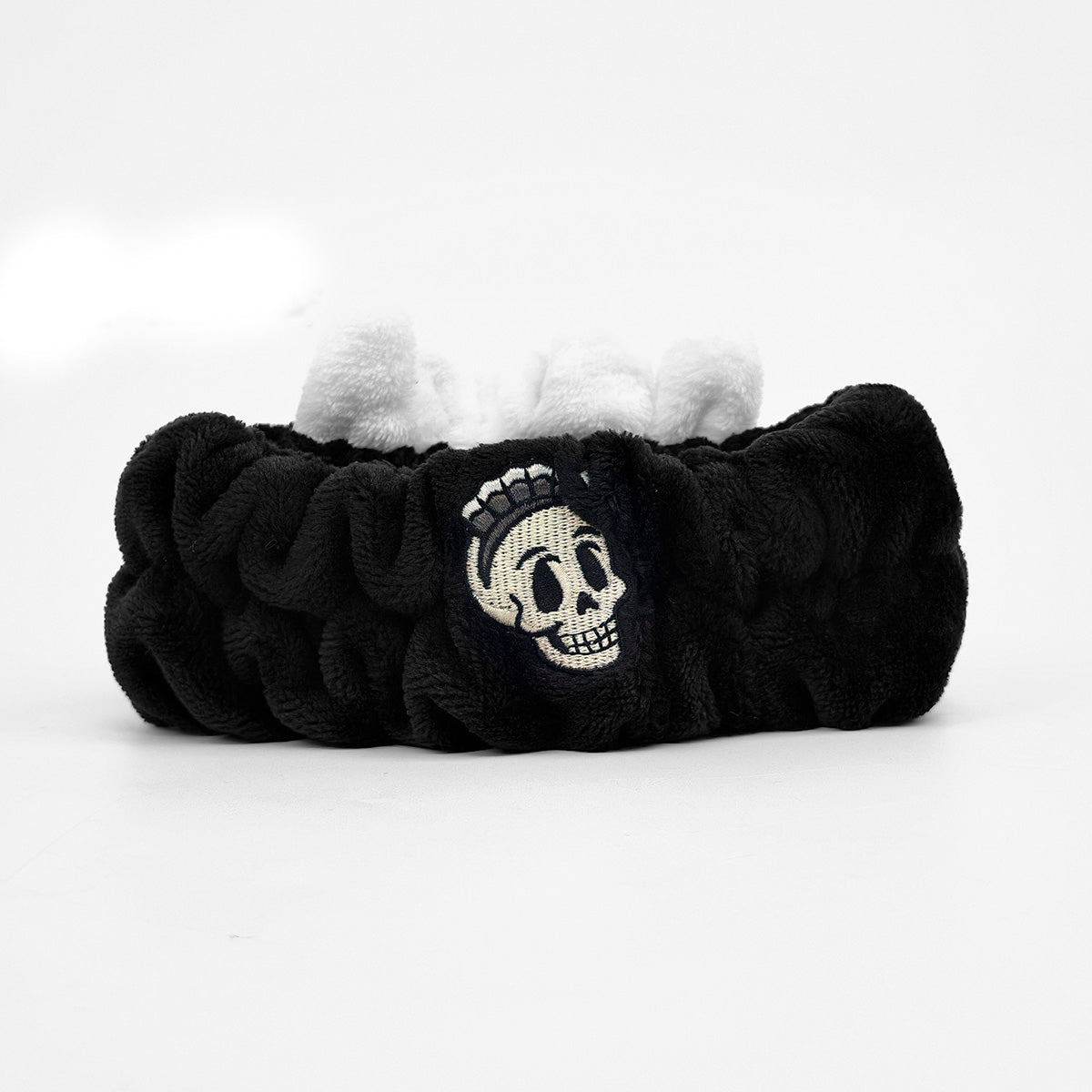 A black plush scrunchie with a white embroidered skull patch on the front, set against a plain white background.