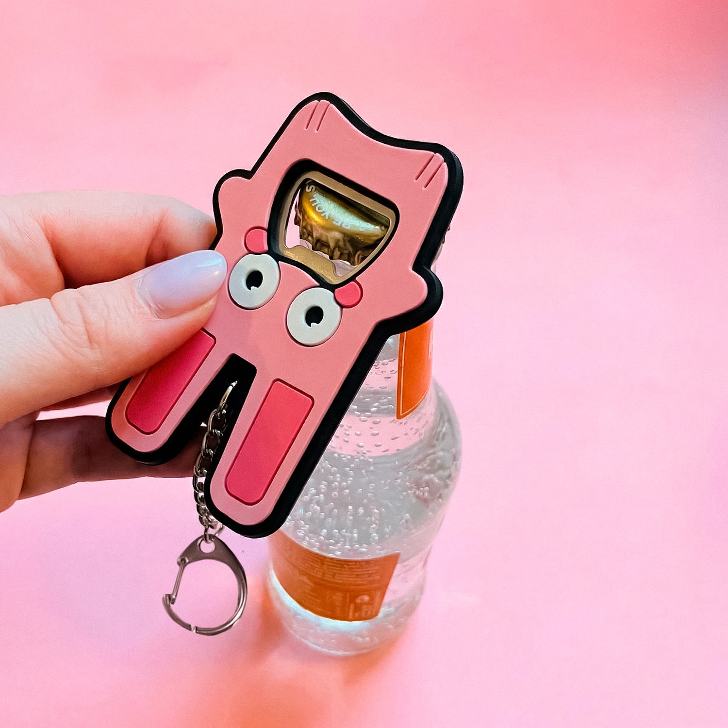 Pink cartoon-style bottle opener with a keychain held over a bottle on a pink background