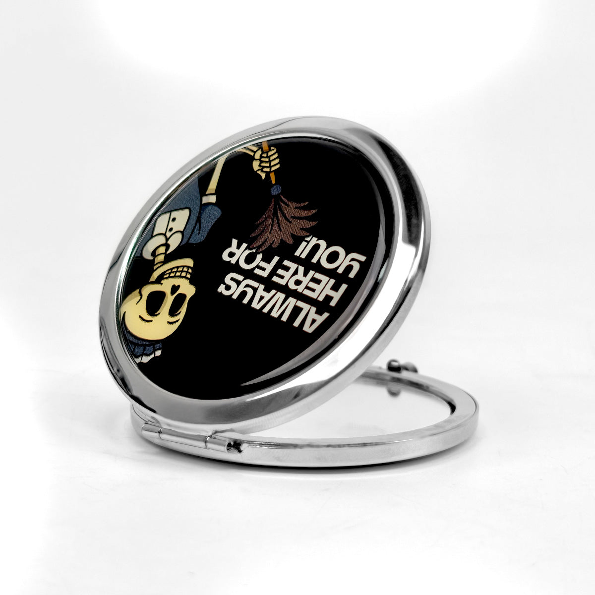 A round compact mirror with a polished silver metal frame and hinge, open to reveal a black reflective surface featuring a cartoon skull character and the text 'ALWAYS HERE FOR YOU!'.