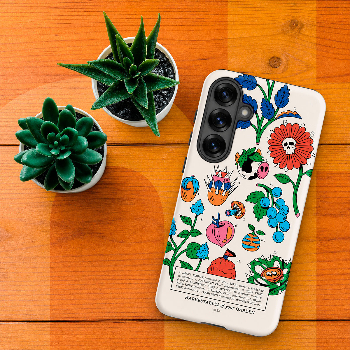 A white phone case with a colorful hand-drawn style illustration of autumn harvest vegetables and flowers, displayed on a wooden table next to two small potted succulents.