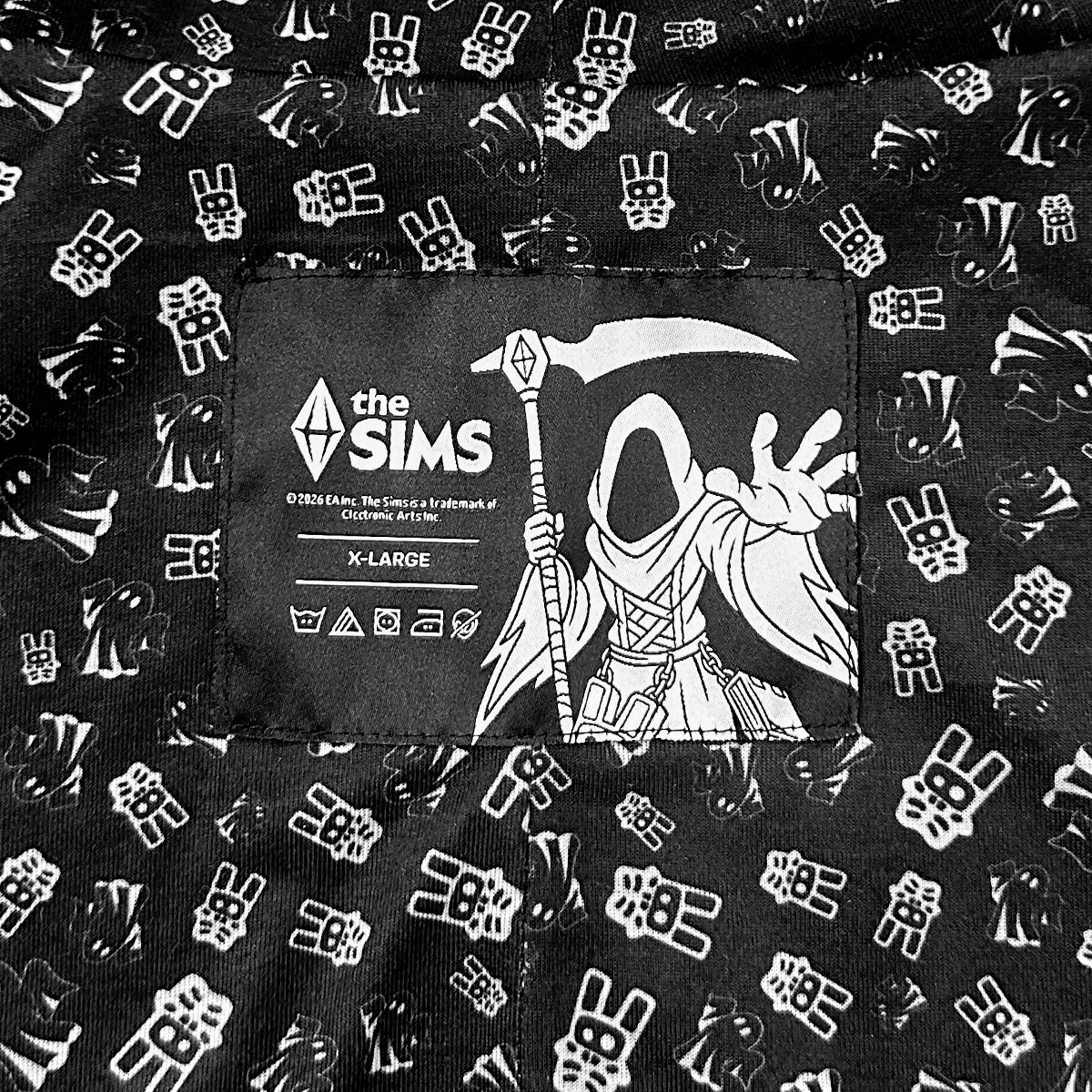 Close-up of a black garment with an allover white repeating bunny-and-scythe pattern and a central sewn label reading 'the SIMS' with a grim reaper illustration and 'X-LARGE'.