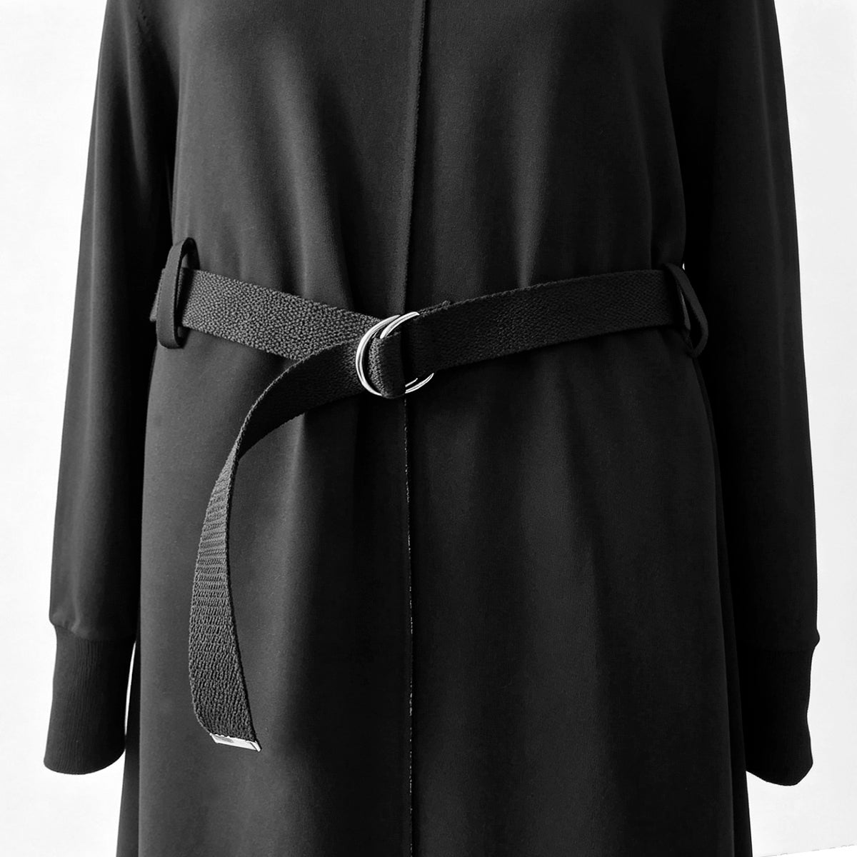 A close-up of a black long-sleeve coat or dress with a matching fabric belt and a silver-tone metal ring buckle, shown against a plain light background.