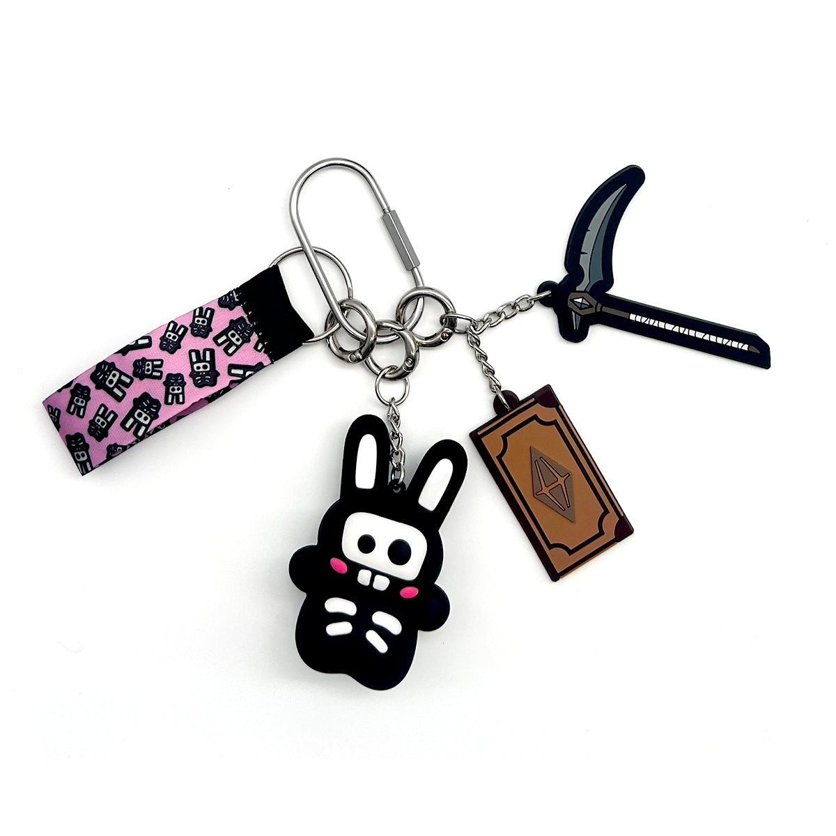 A multi-piece keychain set featuring a black bunny charm with pink cheeks, a brown book charm, a black pickaxe charm, and a pink fabric loop with black cartoon graphics, all attached to a silver split ring and carabiner on a white background.