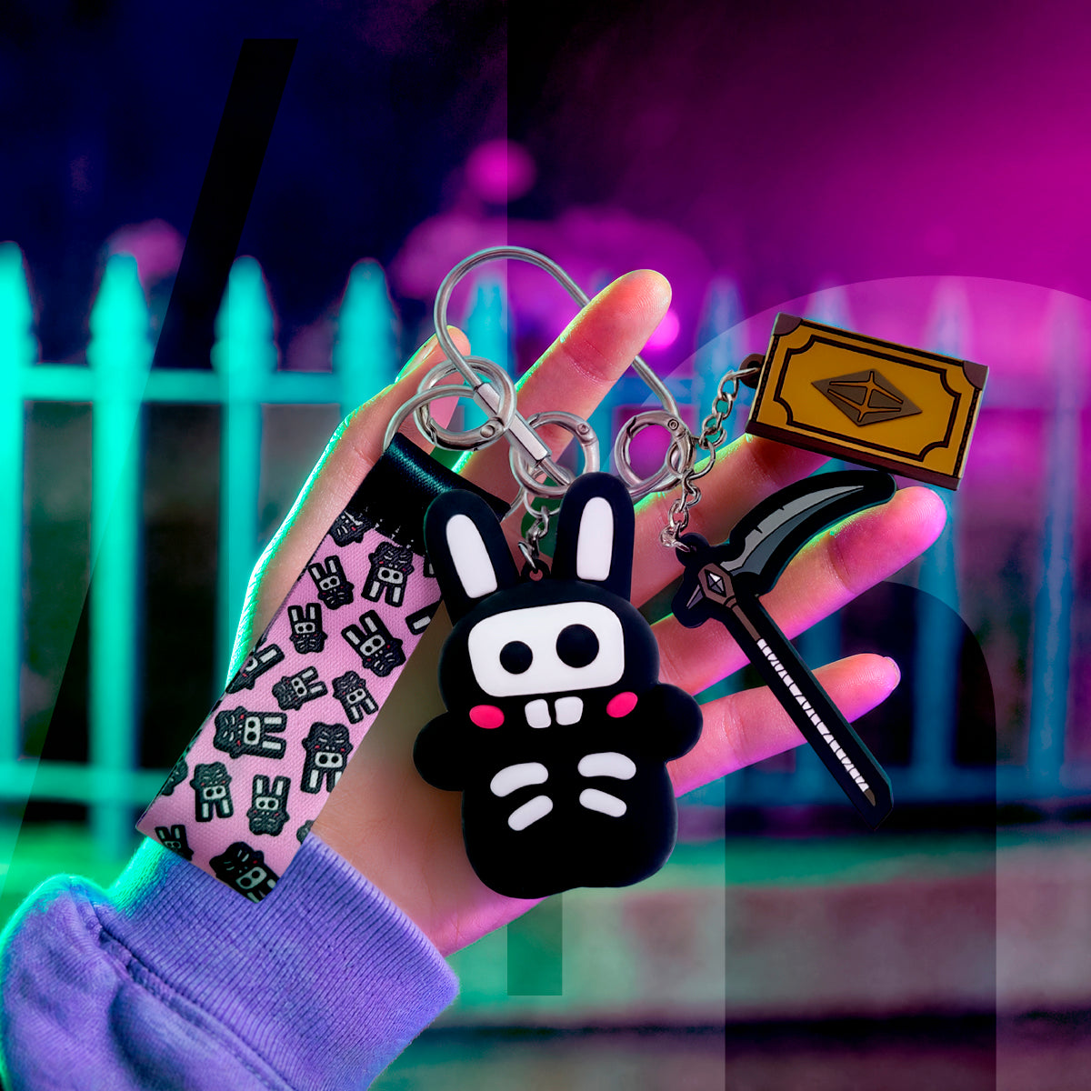 A hand wearing a lavender sleeve holds a set of anime-themed keychains with a black bunny character, a pink patterned wrist strap, a black knife, and a gold ticket, set against a blurred neon-lit background.
