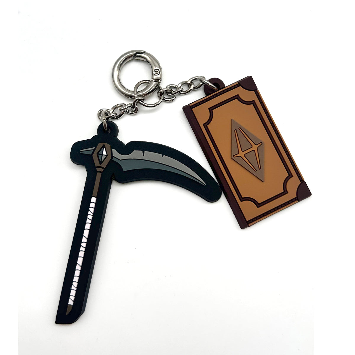 A keychain featuring a black scythe-shaped charm and a brown book-shaped charm with a metallic emblem, attached to a silver metal ring on a white background.