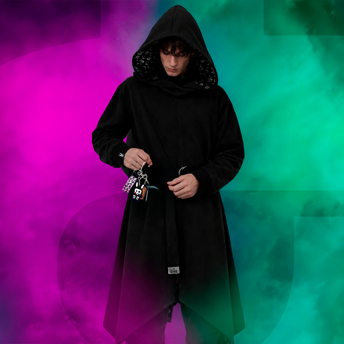 A person wearing a long black hooded robe or coat with a high collar, standing against a smoky multicolor background that shifts from bright pink on the left to teal and green on the right. The person is holding keys in one hand and has a small white tag near the lower front hem.
