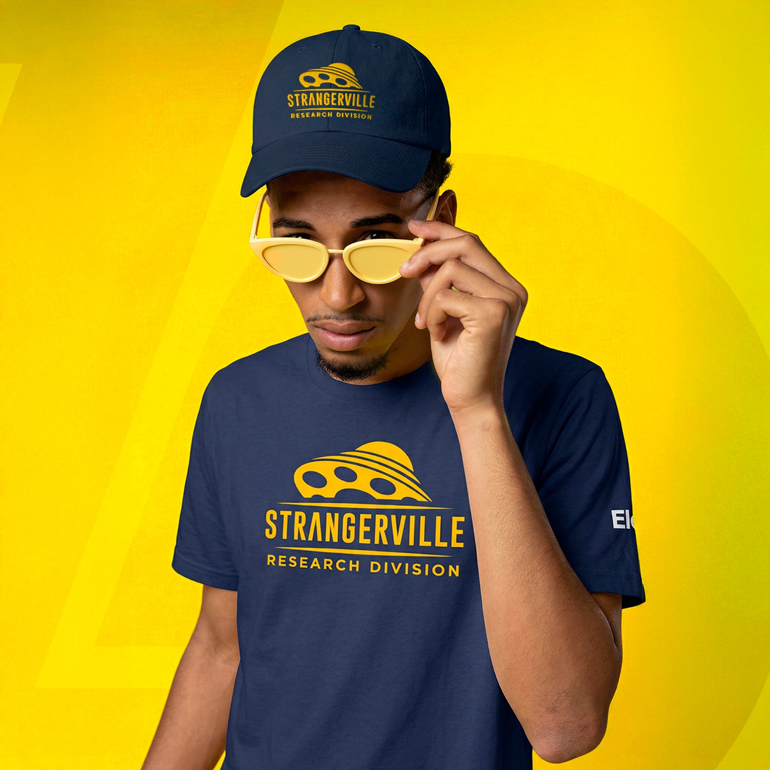 Person wearing a navy 'Strangerville Research Division' t-shirt and cap on a yellow background